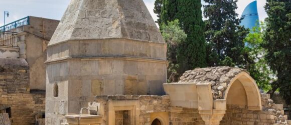 Private custom-built mausoleum with conical roof and archways, reminiscent of Mausoleum Of The Shirvanshah, surrounded by lush trees; contrasted by a modern glass skyscraper in the background. Perfect for high-end clients seeking expert craftsmanship in legacy architecture across the United States.