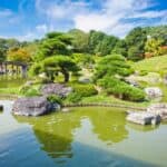 Serene Japanese garden with island, trees, rocks, and pond near Nintoku Mausoleum; distant wooden bridge under clear blue sky.